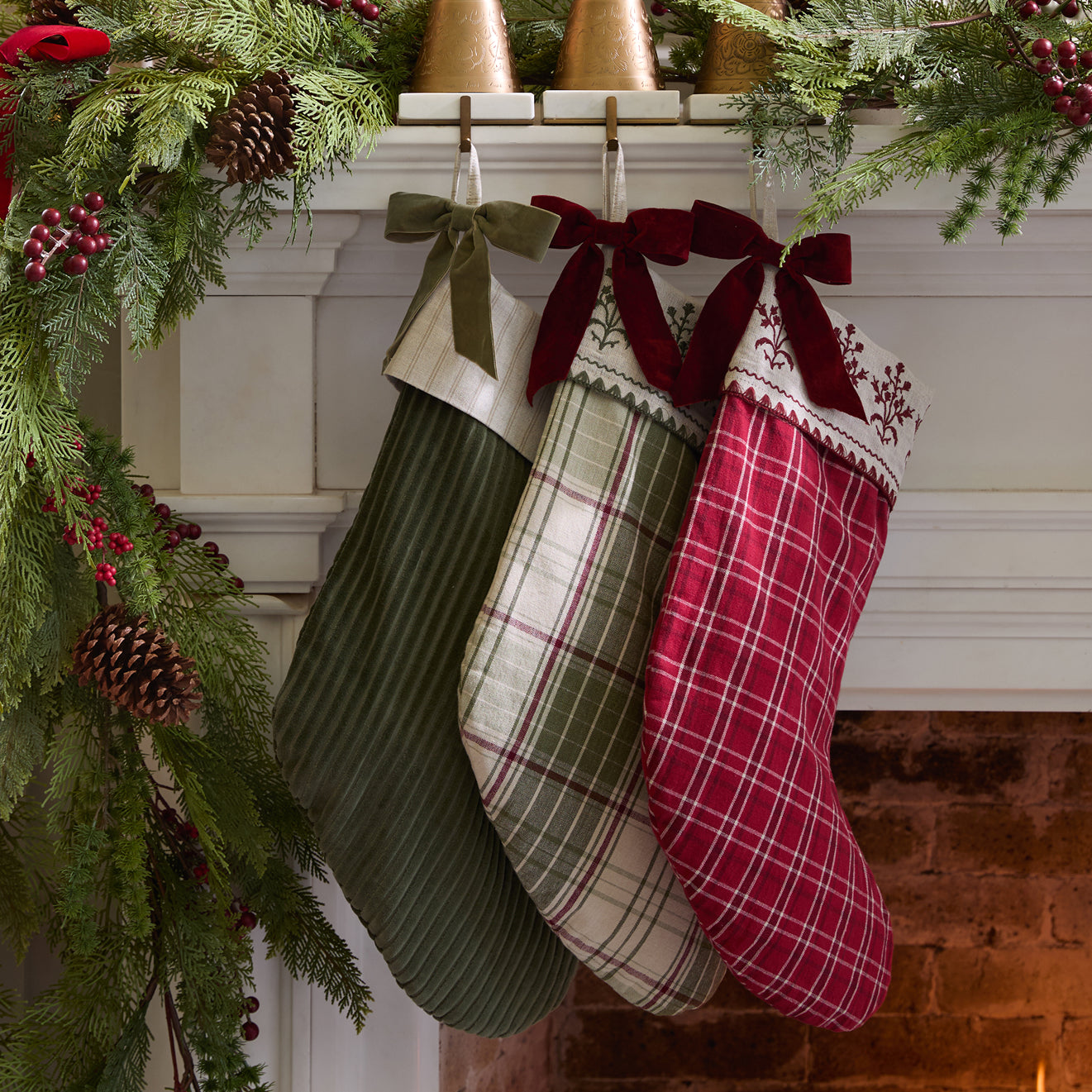 Three Christmas stockings hanging on a fireplace mantle decorated with greenery and pinecones.
