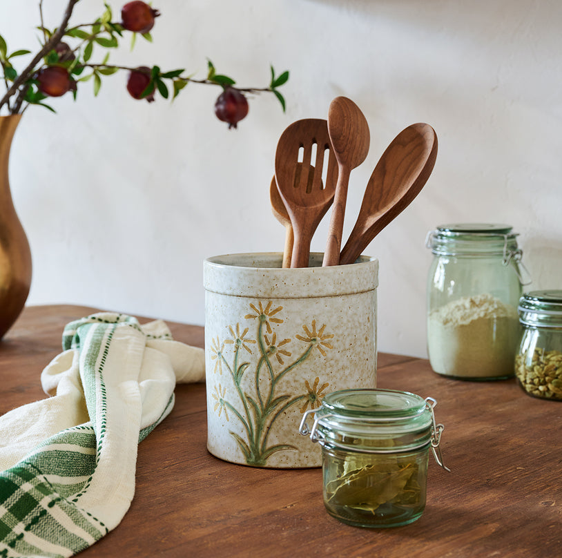 Wooden utensils in a decorative container on a wooden table with jars and a branch.