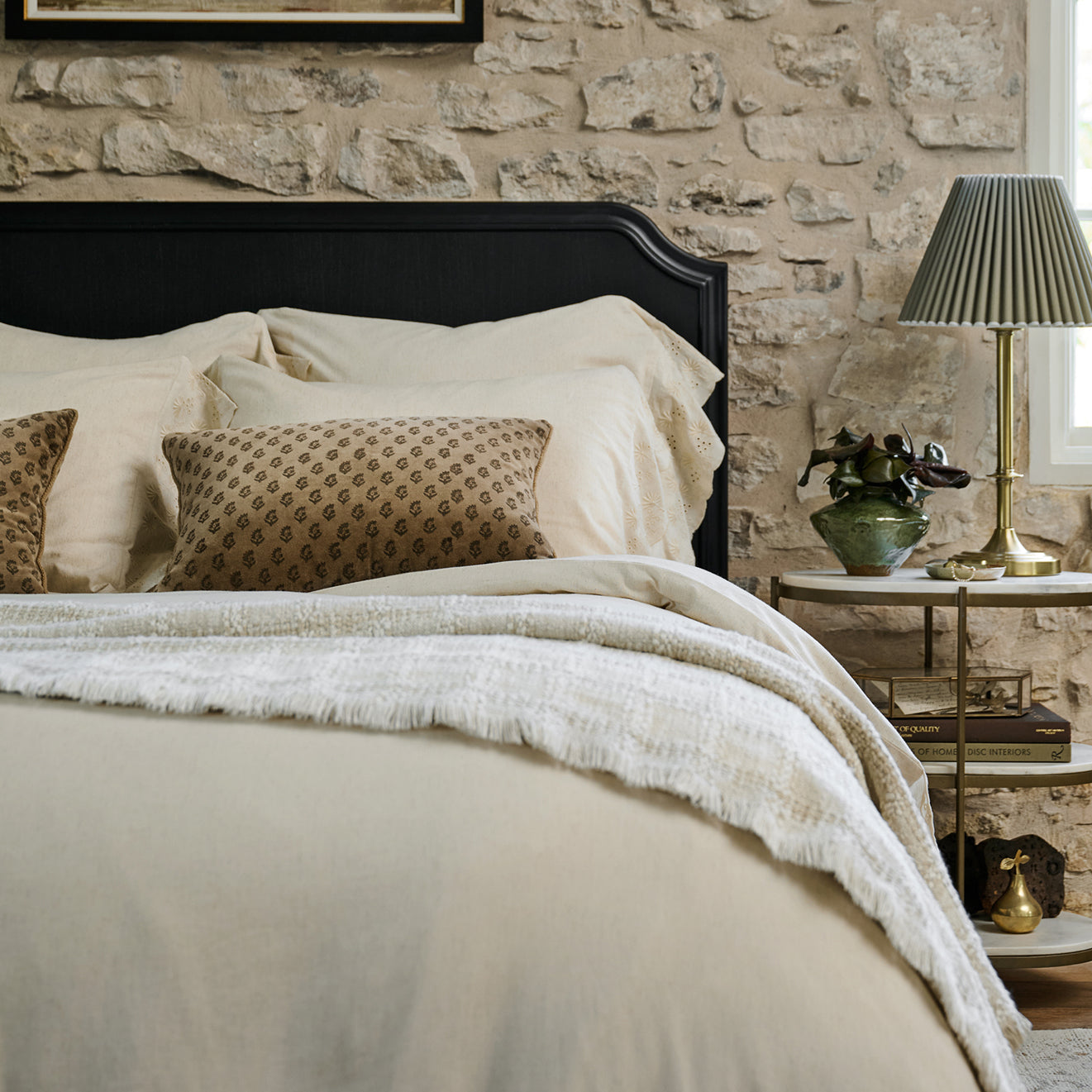 Neatly made bed with decorative pillows against a stone wall, nightstand with lamp and decor items.