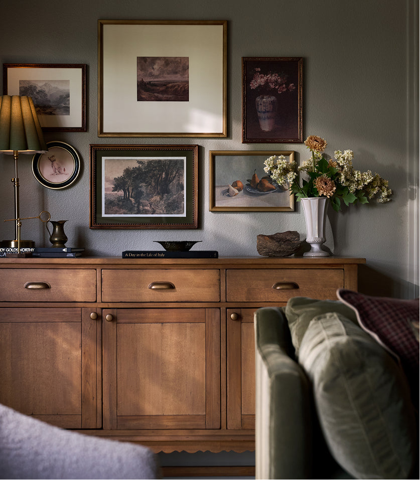 A warm brown wooden sideboard or credenza in a styled room, decorated with framed landscape and still life paintings, a table lamp, and a white vase of flowers. The foreground shows the corner of a green sofa.
