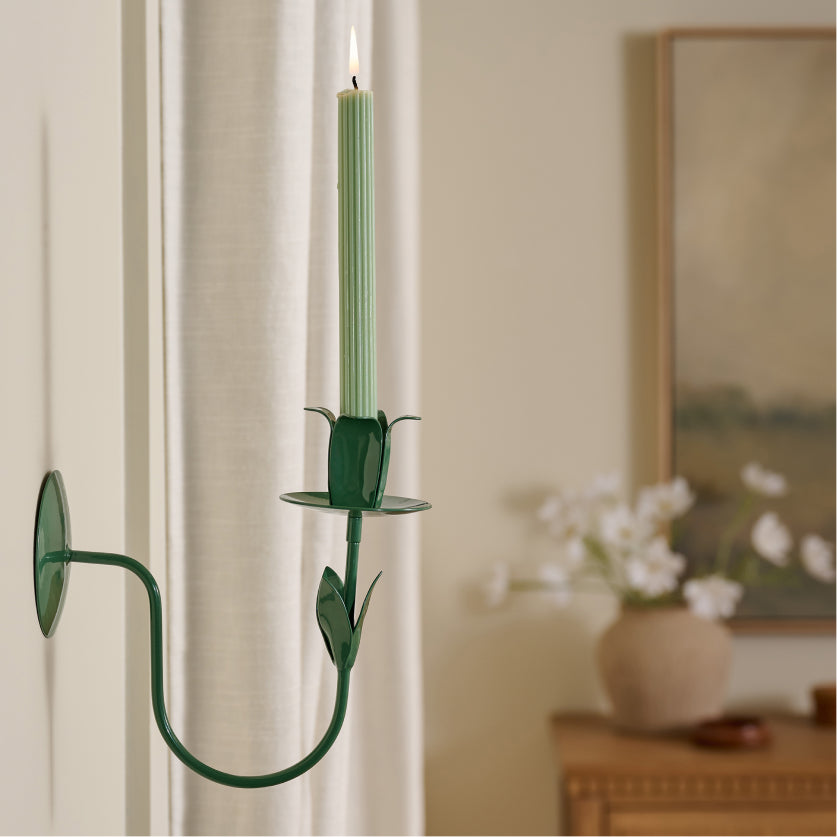 Metal tulip themed green candle holder sconce with a green taper candle on a white wall, with a blurred background of a room.