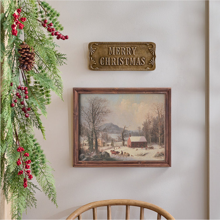 Decorative Christmas scene with a 'Merry Christmas' sign, greenery, and a framed winter landscape painting on a wall.