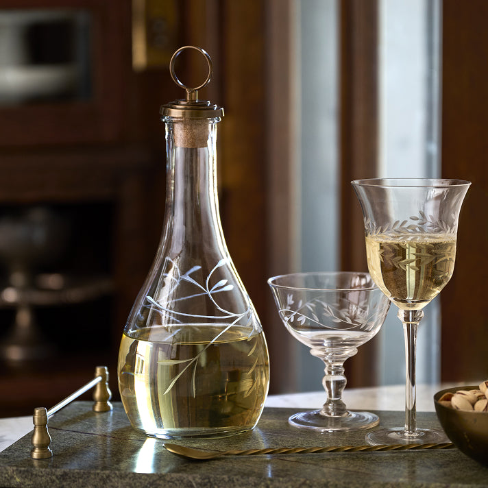 Etched glass decanter and two wine glasses sitting on a marble tray with a twisted brass spoon.
