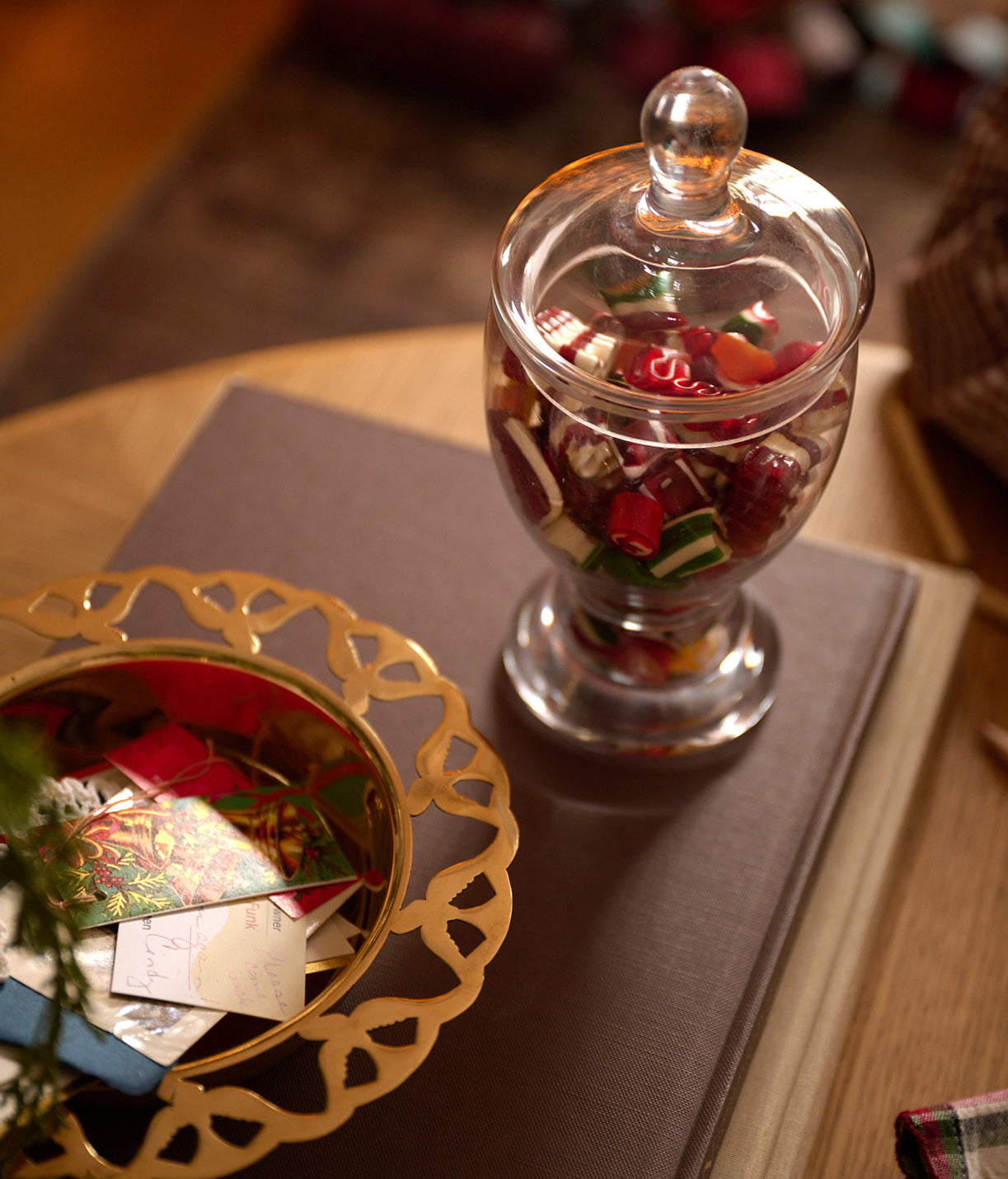 A clear glass apothecary-style jar with a round glass lid, filled with colorful candy pieces, placed on a brown book next to a gold decorative bowl containing small cards.
