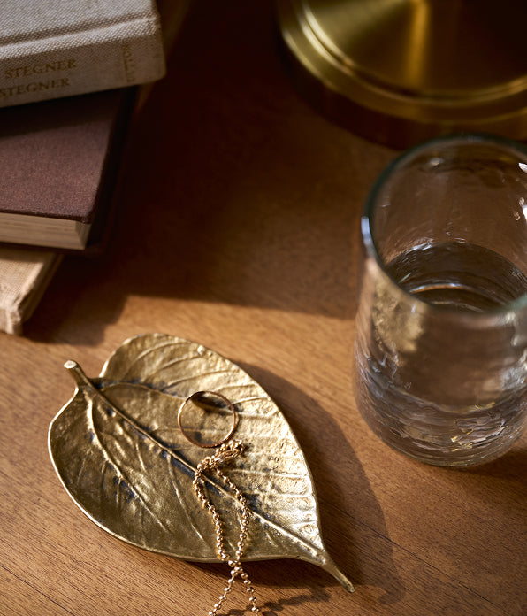 A gold-toned leaf-shaped jewelry stand holding a ring and chain, placed on a wooden tabletop beside a glass of water and stacked books.