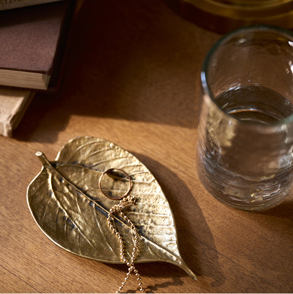 A gold leaf-shaped jewelry organizer and tray holding a gold ring and chain, styled on a wooden table next to a glass of water.