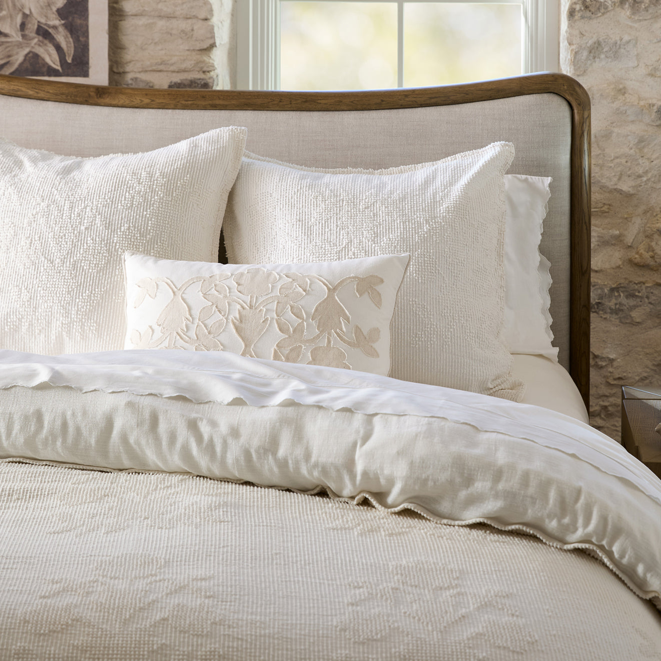 Neatly made bed with white bedding and decorative pillows against a stone wall.