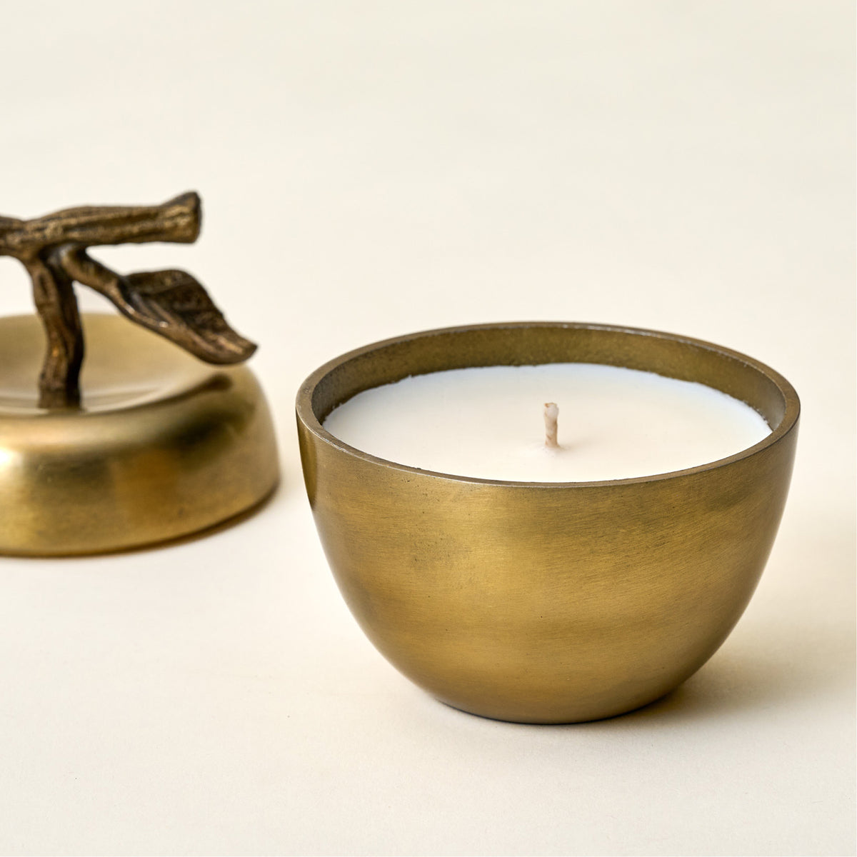 A brass candle holder with a white wax candle and a single wick, shown on a light beige background with its matching brass lid and leaf-shaped handle partially visible in the background.