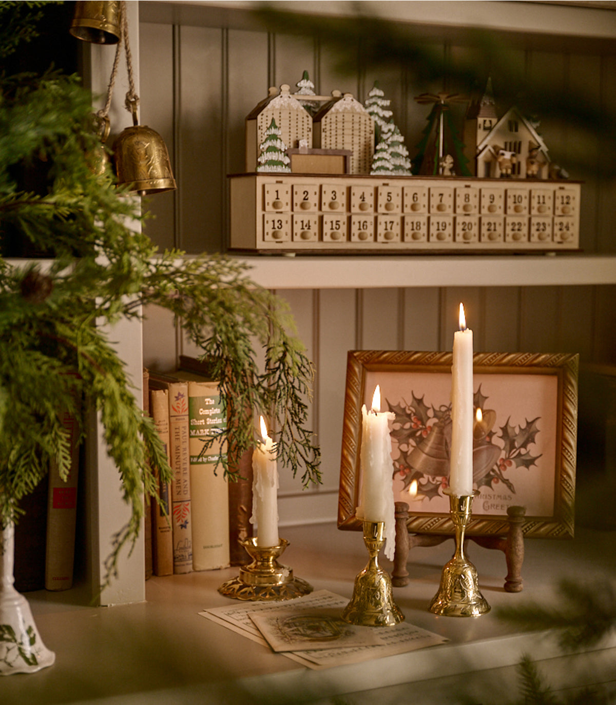 A cozy holiday-themed shelf display featuring three lit white taper candles in ornate gold-toned holders, a framed holly print, books, greenery, and a wooden advent calendar on a paneled wall.