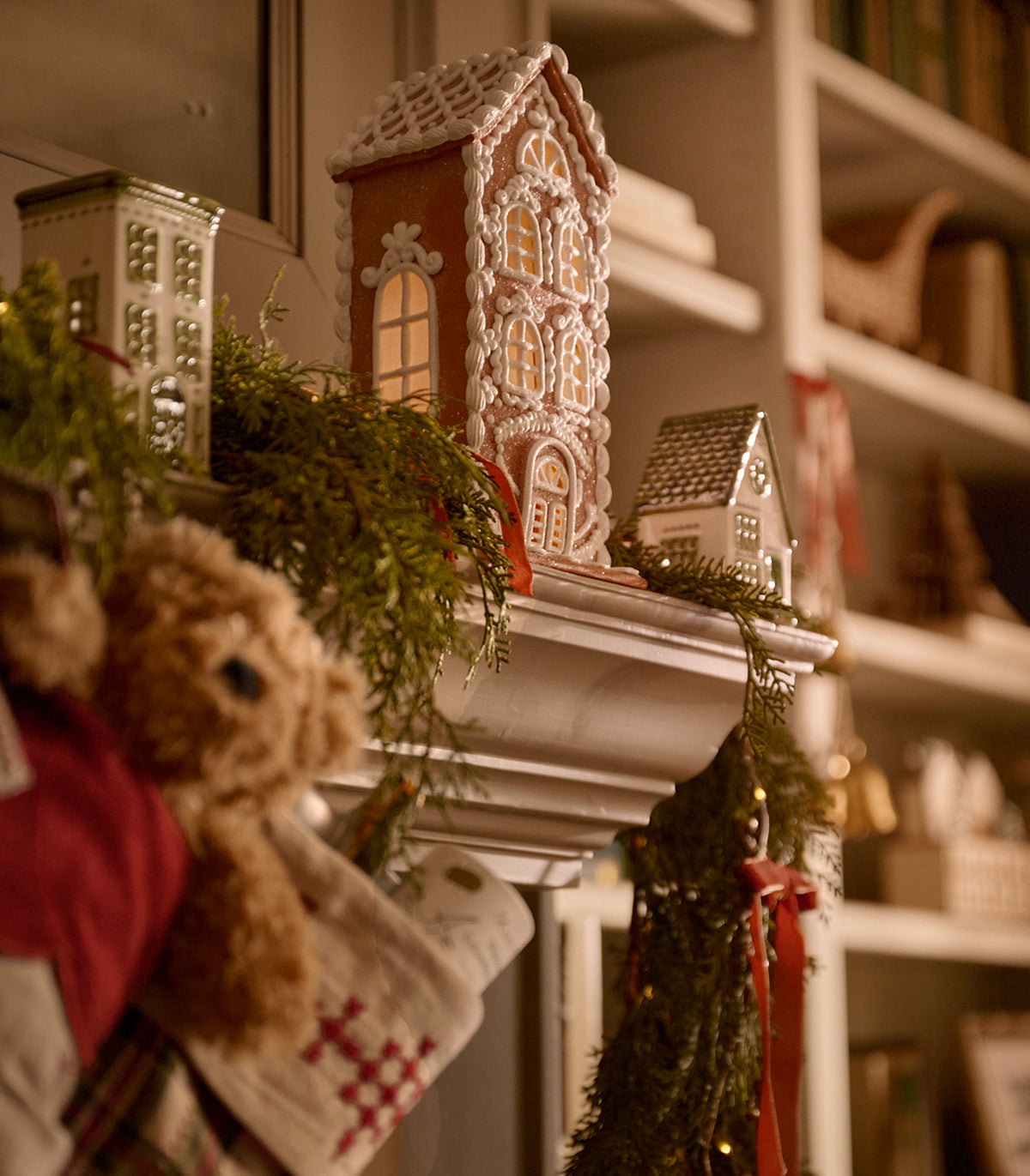 A decorated holiday mantel with gingerbread house ornaments, green garland, stockings, and a teddy bear, set against a softly blurred bookshelf background.