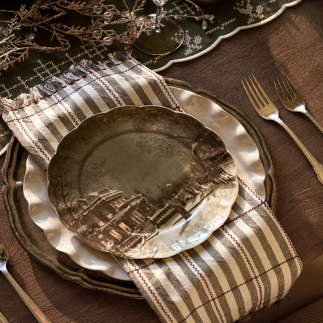 A place setting featuring a rustic ceramic dinner plate with a detailed cityscape illustration, layered over a striped beige and brown napkin and a matching charger plate on a dark wood table.