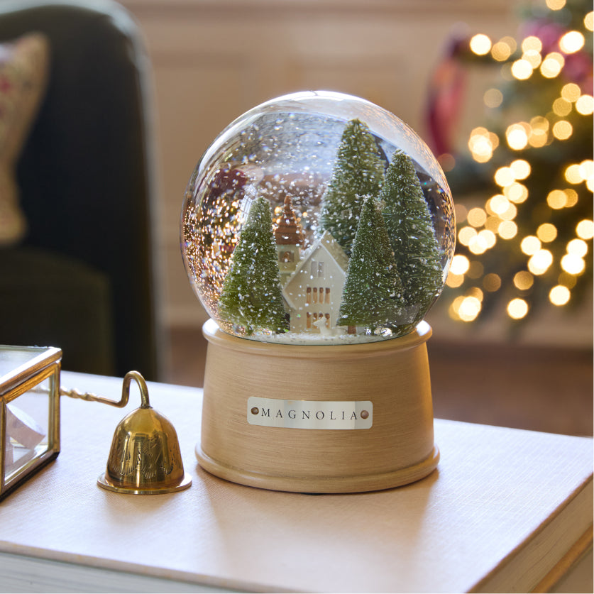 Snow globe with the Silos chapel and pine trees, on a table with blurred Christmas lights in the background.
