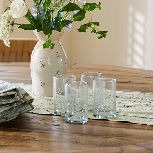 Clear glass tumblers on a wooden table with a white vase containing flowers.
