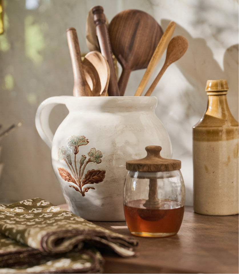 A rustic white ceramic pitcher decorated with a botanical floral pattern, filled with wooden spoons and utensils, set on a wooden table alongside a glass honey jar and a stoneware bottle.
