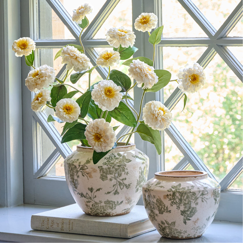 Two decorative distressed vases with floral patterns on a windowsill with a flower arrangement.