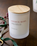 Frosted candle with wooden lid labeled 'Joyeux Noel' on a wooden surface with decorative elements.