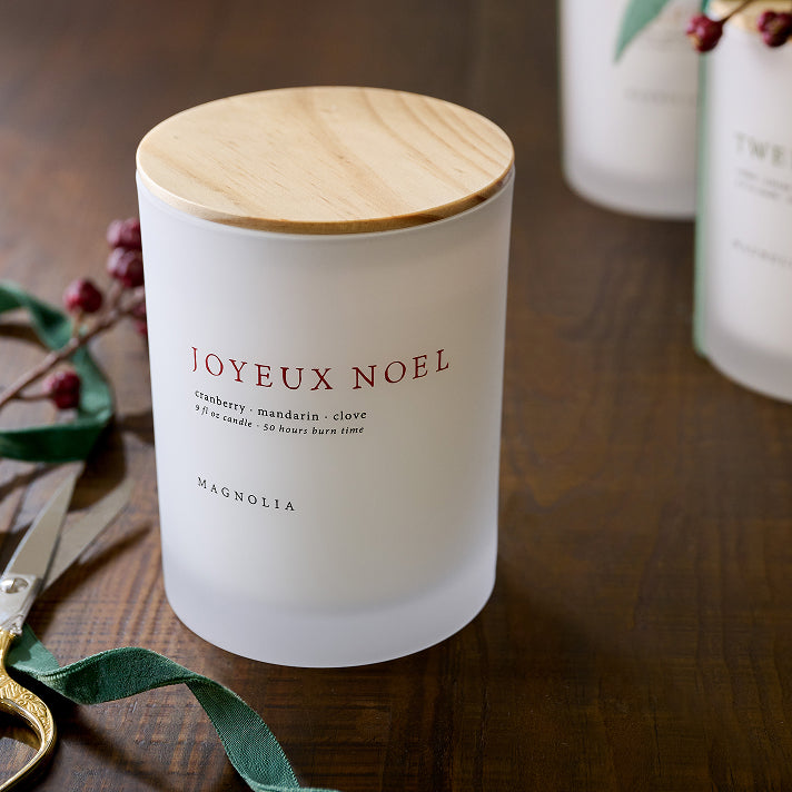 Frosted candle with wooden lid labeled 'Joyeux Noel' on a wooden surface with decorative elements.