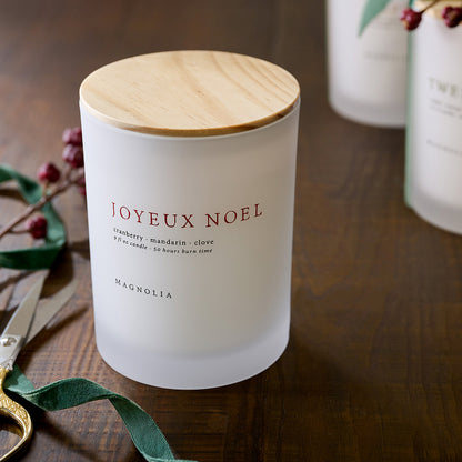 Frosted candle with wooden lid labeled 'Joyeux Noel' on a wooden surface with decorative elements.