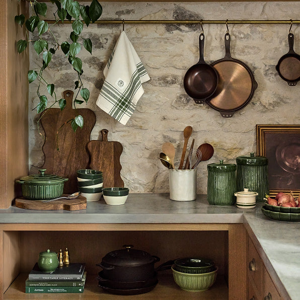 Kitchen counter with green ceramic items, wooden cutting boards, and copper pans against a stone wall.
