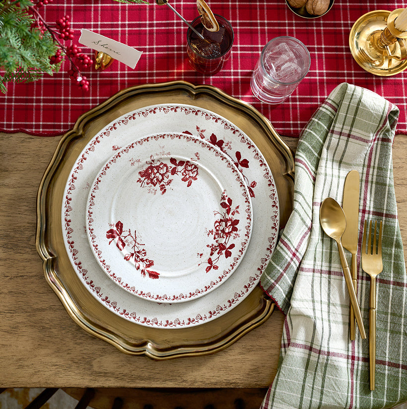 Dining table setting with a red and white floral plate, gold charger, and silverware on a red plaid tablecloth.