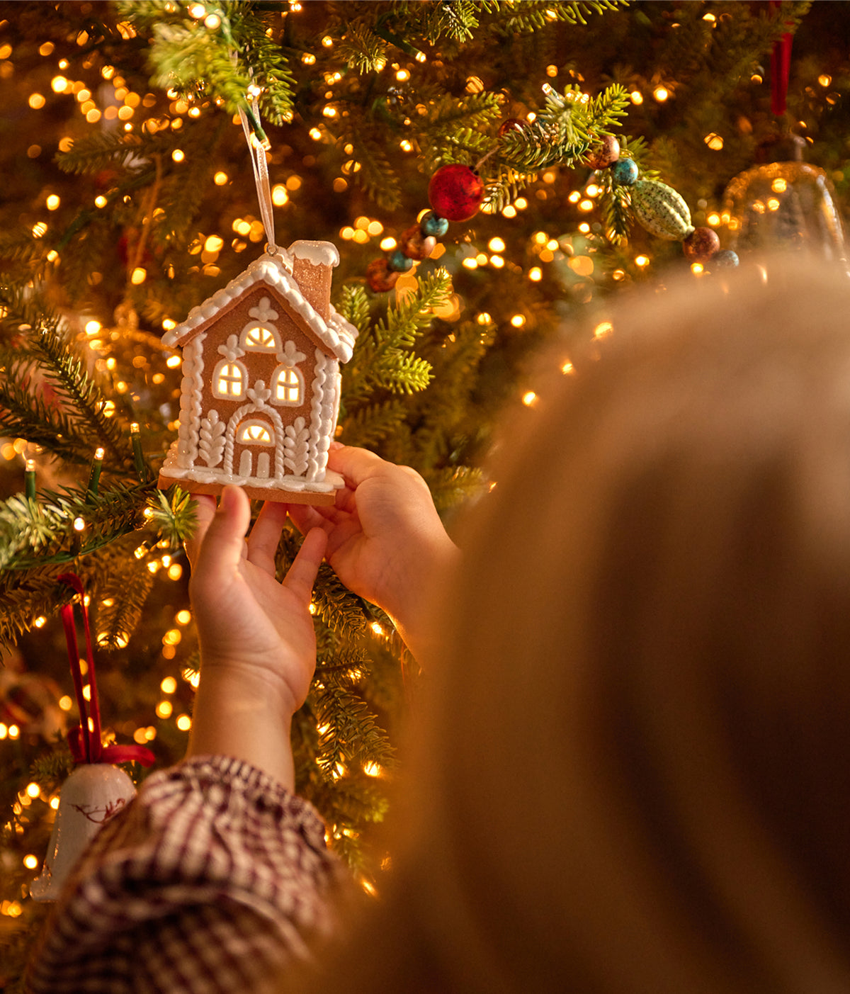 A person hanging a small gingerbread house Christmas ornament on a decorated Christmas tree with warm glowing lights.