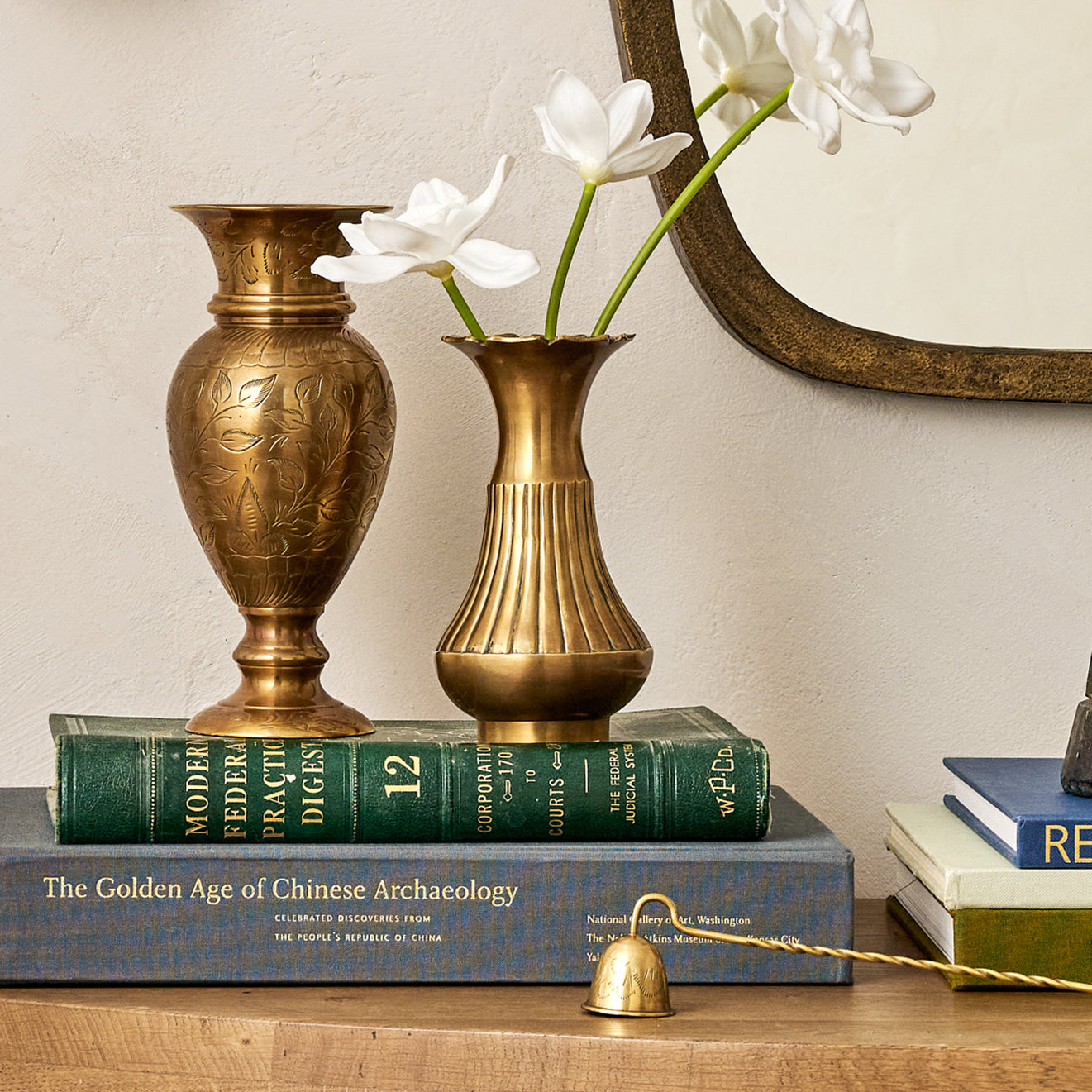 Two brass vases with white flowers on books against a neutral wall.