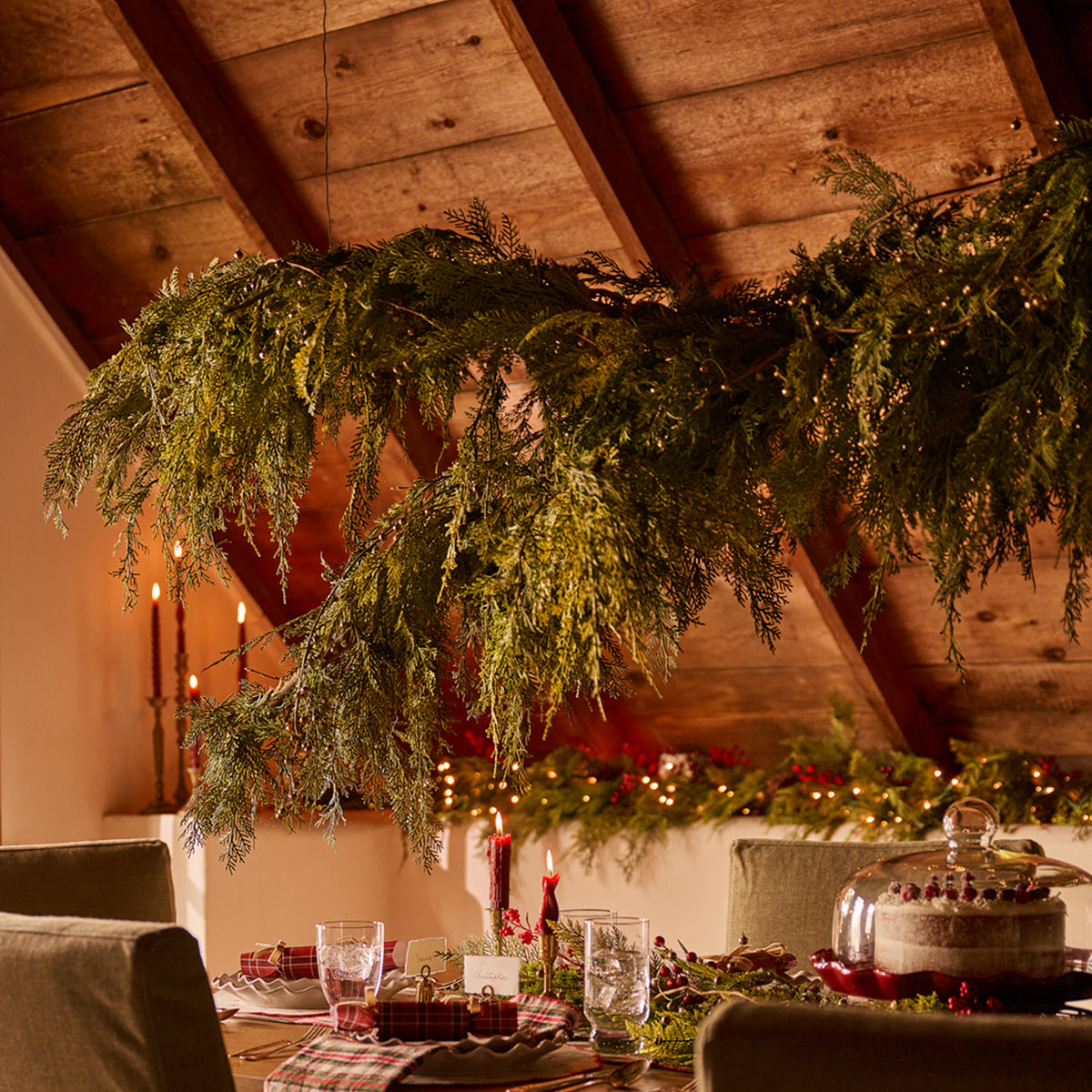 A festive dining table decorated with green garlands hanging from a rustic wooden ceiling, with candles, plates, and a covered cake in a warm holiday setting.