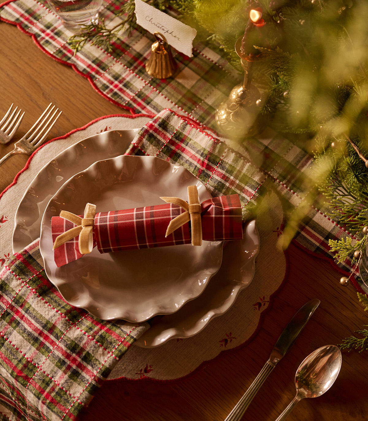 A festive holiday table setting featuring a red and green plaid napkin tied with a beige ribbon, layered on scalloped white plates over a beige placemat. The setting includes a matching plaid runner, silver cutlery, and warm holiday decorations on a wooden table.