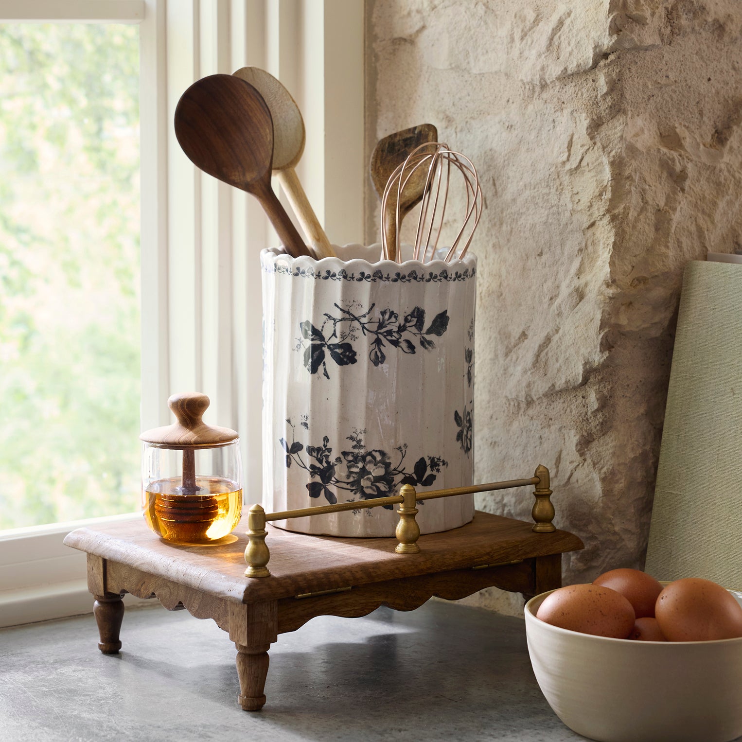Kitchen utensils in a decorative container on a wooden stand with a bowl of eggs in the foreground.
