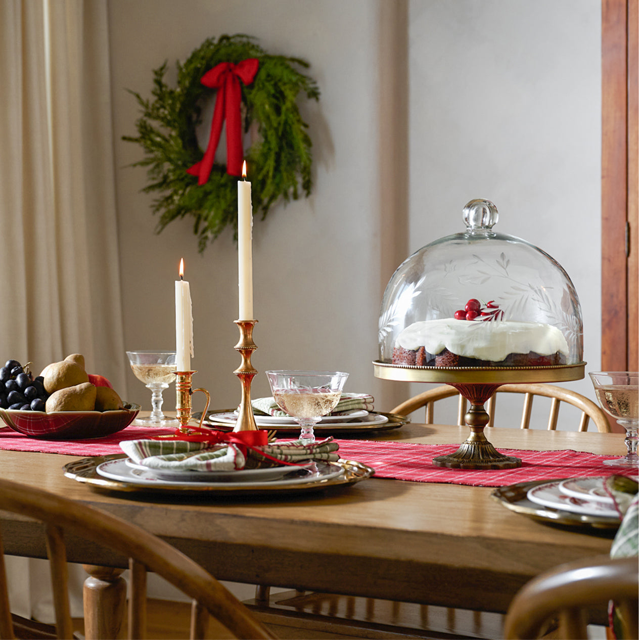 holiday dining table scene with a etched domed cake stand on top a red table runner.  holiday place settings also adorn the table along with two lit taper candles.