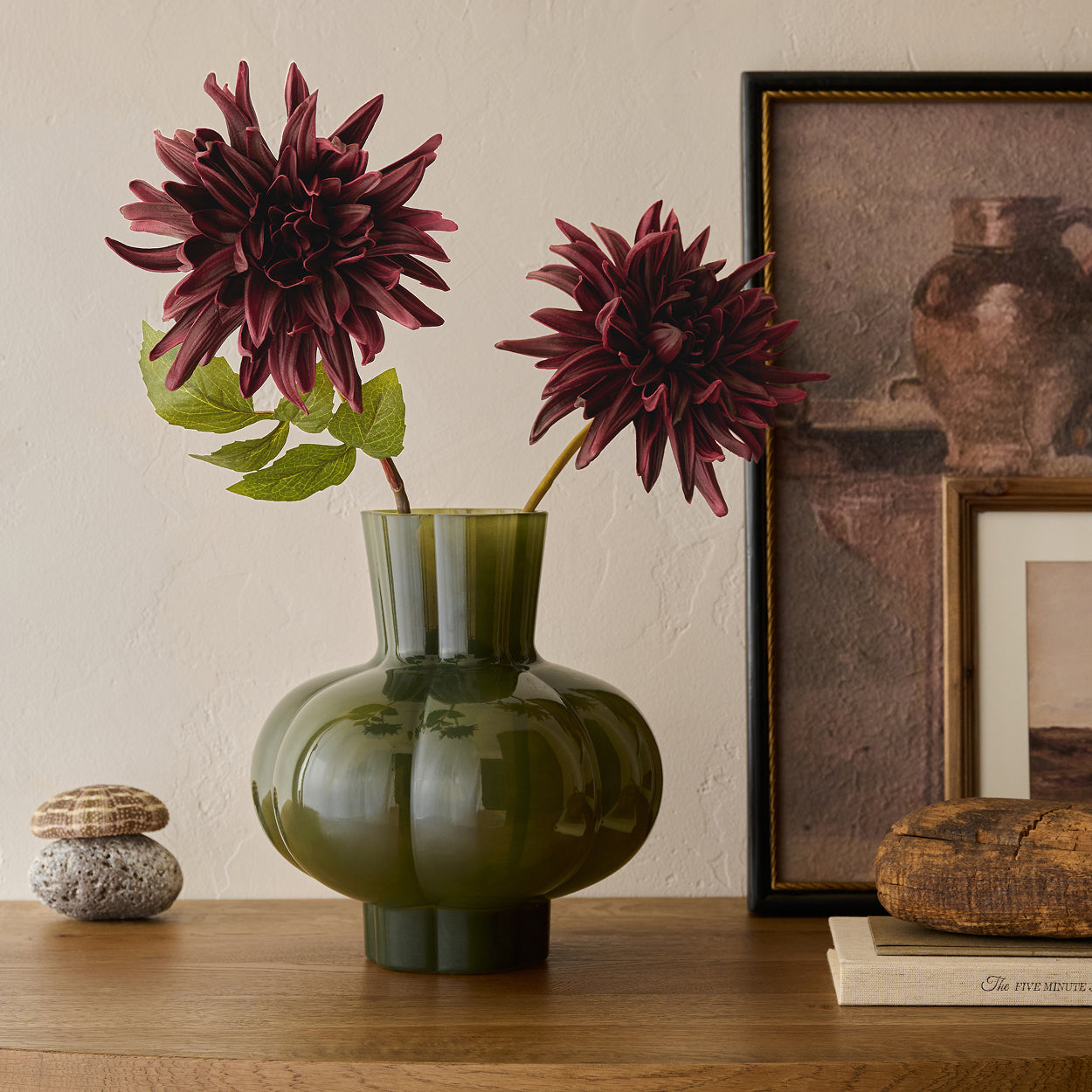 Green vase with burgundy flowers on a wooden surface next to framed artwork.