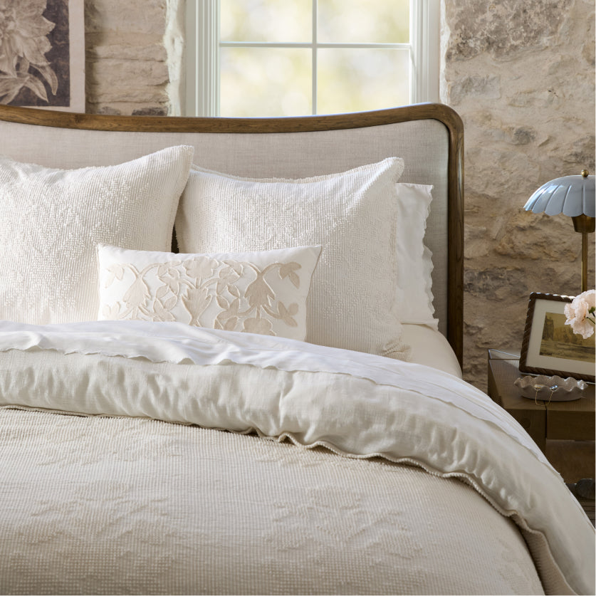 Neatly made bed with white bedding and pillows in a room with stone walls and a window.