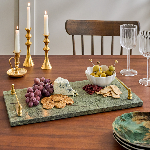 Dining table with a marble cheeseboard featuring grapes, blue cheese, and crackers, accompanied by candles and wine glasses.