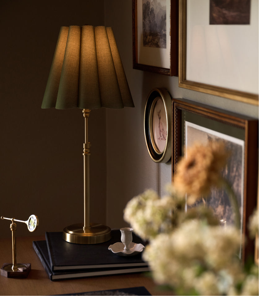 A classic table lamp with a pleated olive green fabric shade and a polished brass base, styled on a dark wooden table next to books and a teacup in a warm, dimly lit room.