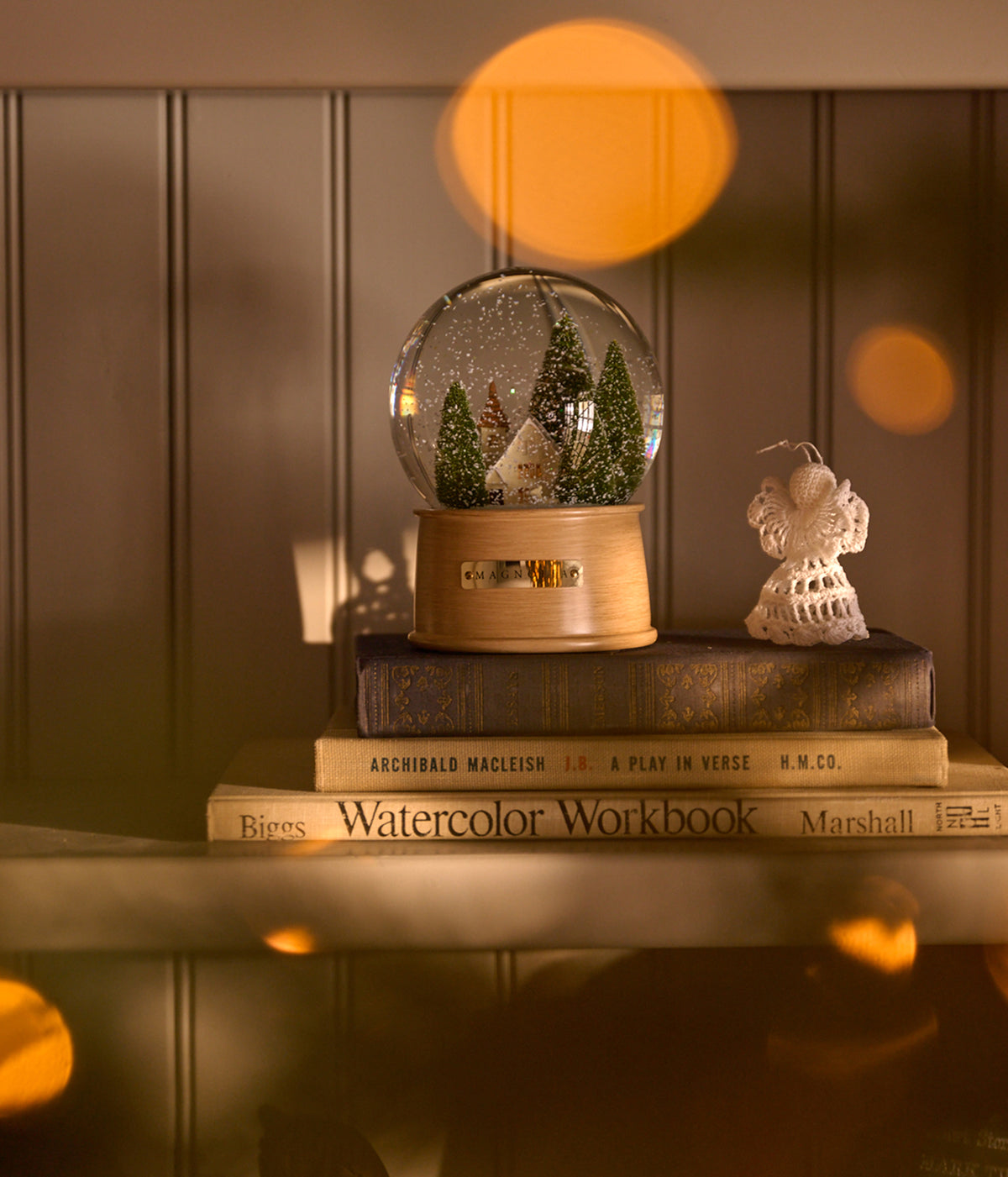 A decorative glass snow globe with a miniature Christmas tree scene and a light wood base sits on top of stacked books, alongside a small white angel figurine, against a warm wood-paneled background with soft orange bokeh lights.