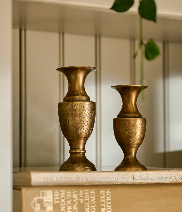 Two ornate brass vases with engraved patterns, displayed on a shelf next to a book.