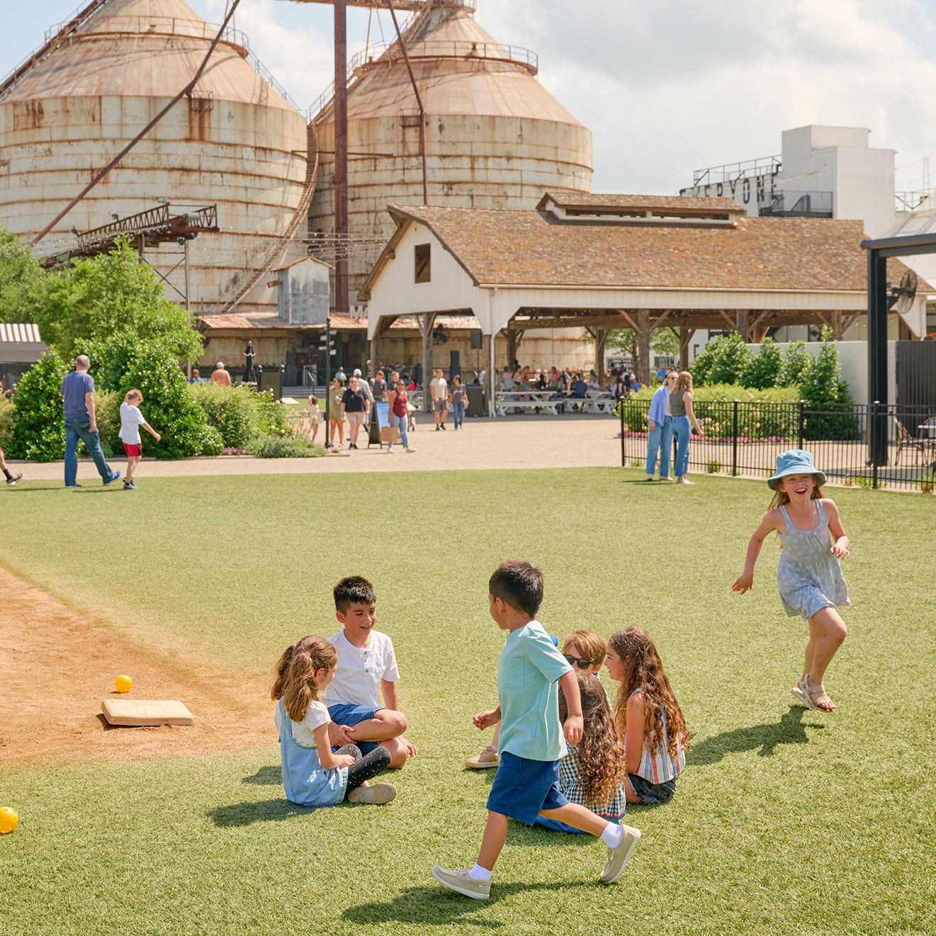 Children playing on a grassy area with large silos and a building in the background.
