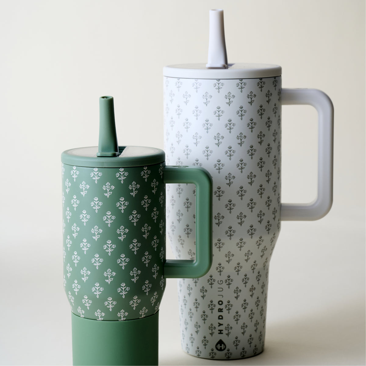 Two Hydrodug handled tumblers with lids and straws, shown on a clean off-white background. One tumbler is sage green and the other is white, both featuring a repeating gray floral pattern.