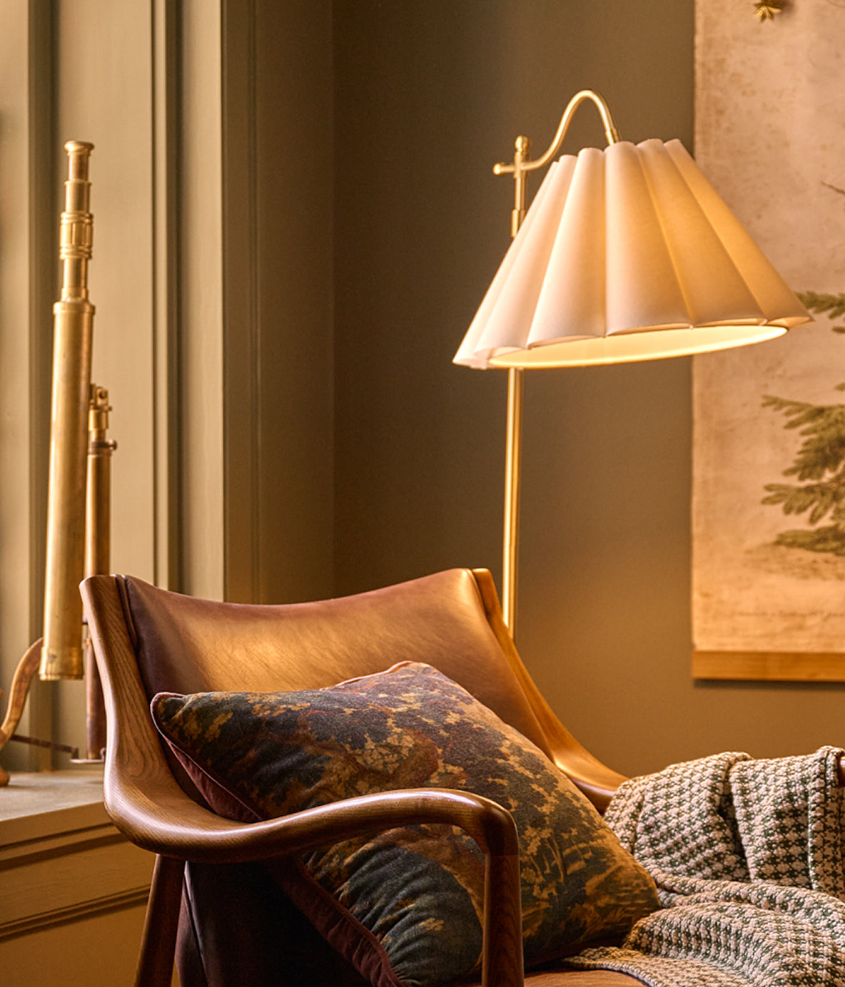 A warm, elegant interior scene featuring a brass floor lamp with a pleated white shade next to a brown leather armchair with a patterned throw pillow and a houndstooth blanket.