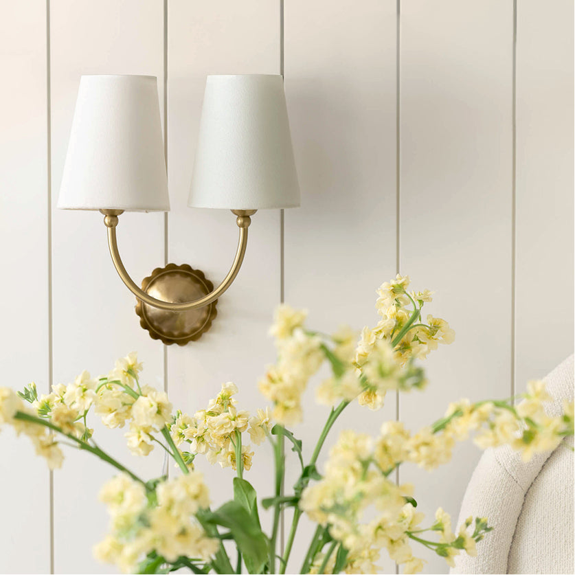 Decorative wall sconce with white lampshades and yellow flowers in the foreground against a light wooden panel background.