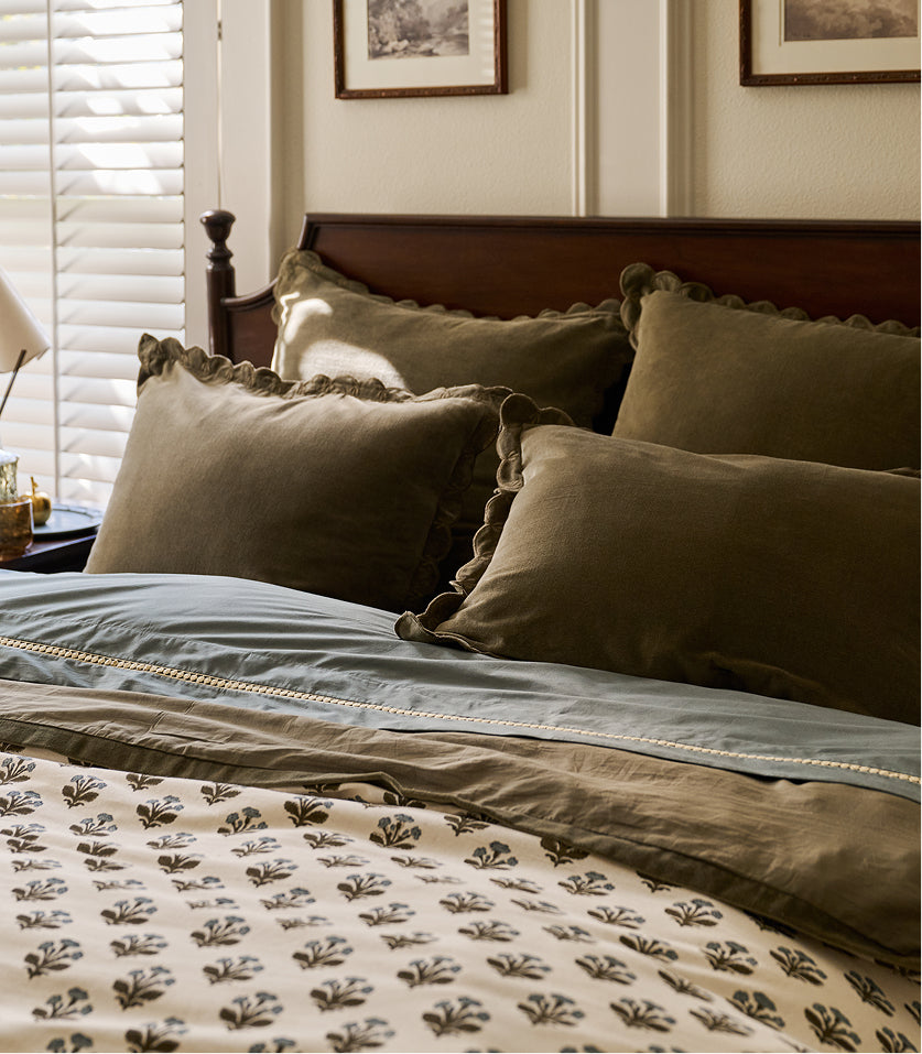 A styled bed in a bedroom setting featuring olive green velvet pillows with scalloped edges, a dusty blue duvet, and a cream-colored comforter with a dark botanical leaf pattern.