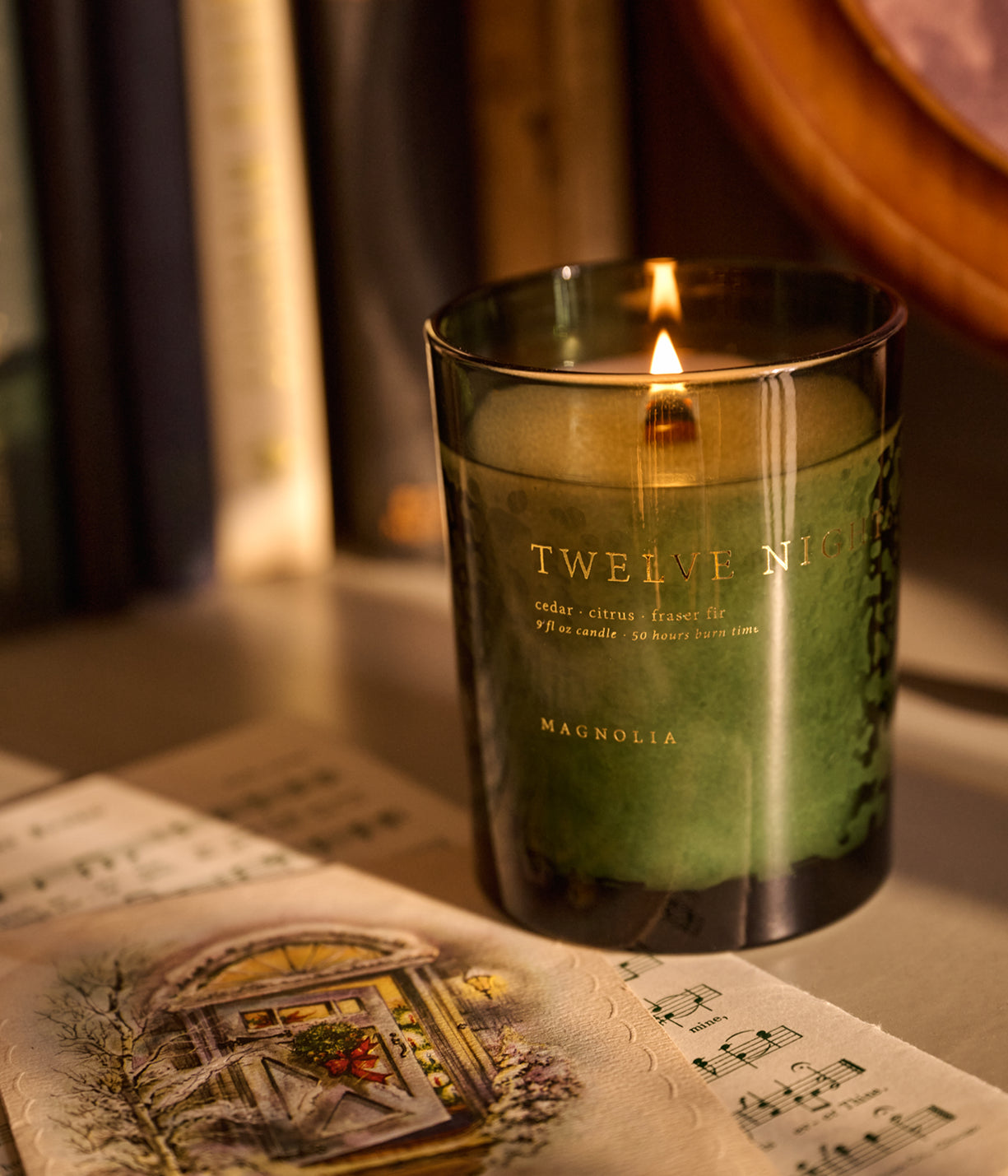 A lit Twelve Nights Magnolia candle in a dark green glass jar, styled on a tabletop next to an illustrated card with a warm, cozy background.