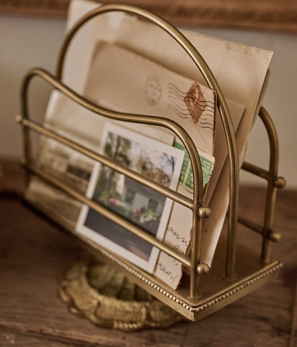 A vintage-style gold metal newspaper or mailer rack holding old envelopes and postcards on a wooden surface.