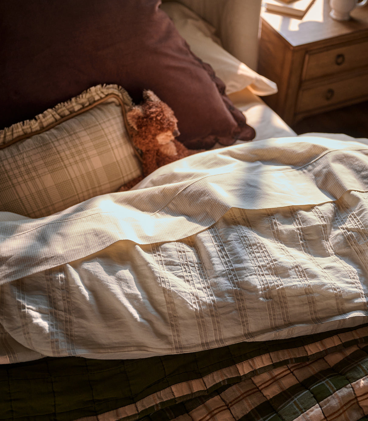 A close-up of a textured cream quilted duvet cover on a bed, styled with a plaid accent pillow, a dark brown pillow, and a small teddy bear, in warm natural sunlight.