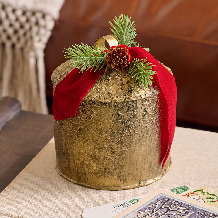 Decorative candle with gold bell, red ribbon and greenery on a table.