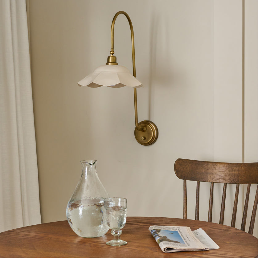 Wall-mounted lamp with a glass shade above a wooden table with a carafe, glass, and newspaper.