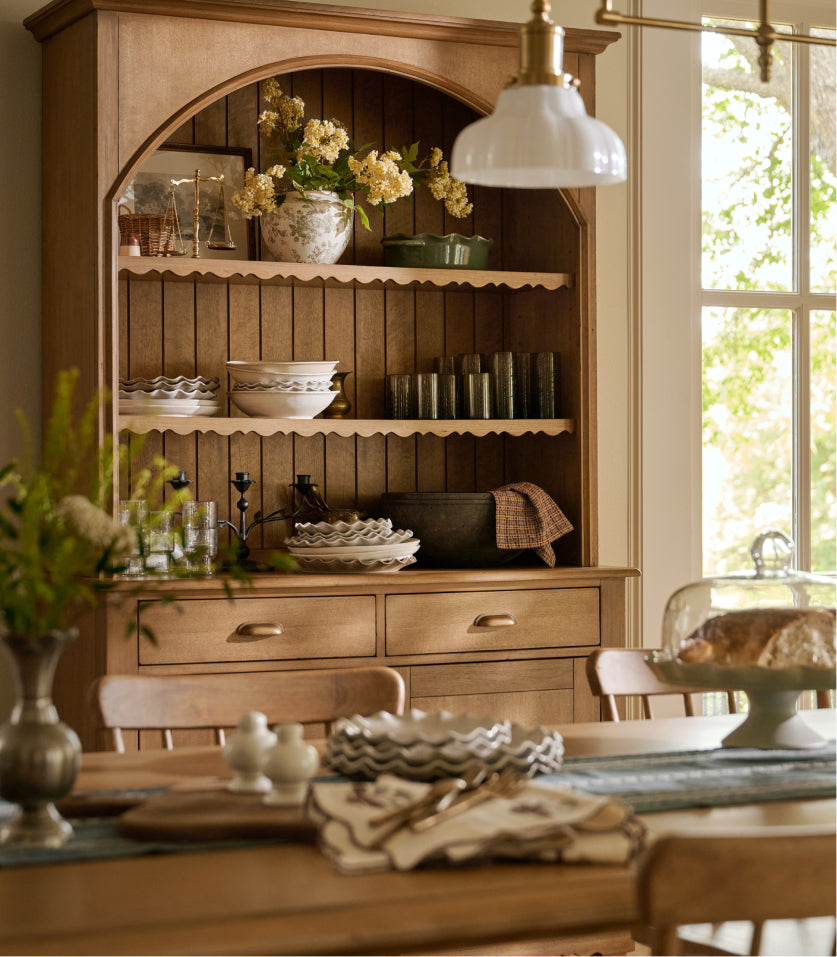 A warm wood hutch or china cabinet in a bright dining room, styled with white dishes, floral arrangements, glassware, and a hanging white dome lamp.