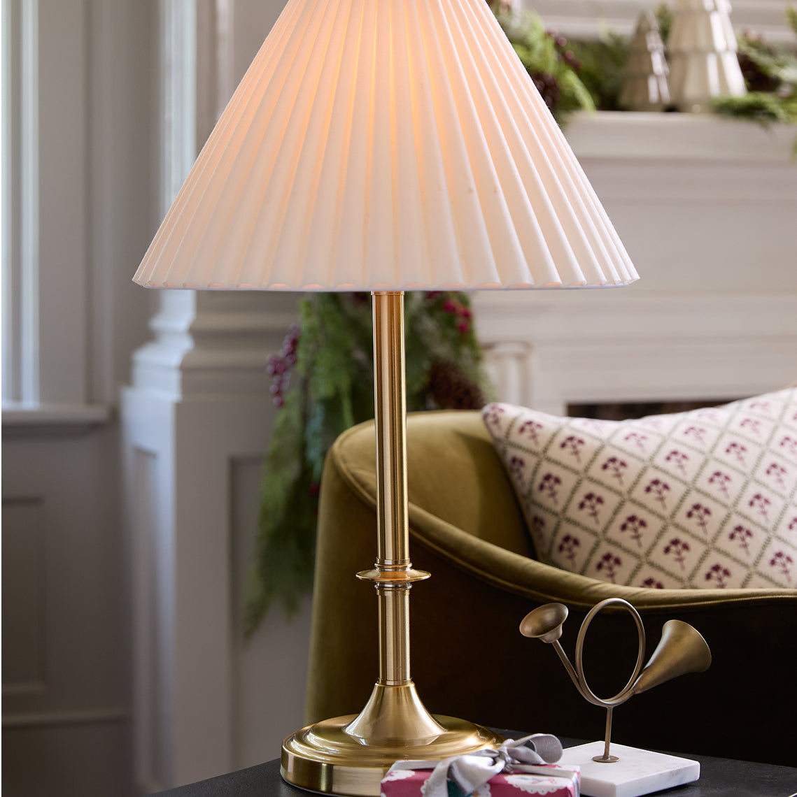 Gold table lamp with pleated white shade on a surface, with a decorative chair and greenery in the background.