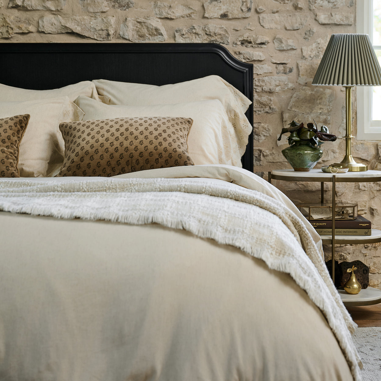 Neatly made bed with beige bedding against a stone wall, with a lamp and decorative items on a nightstand.