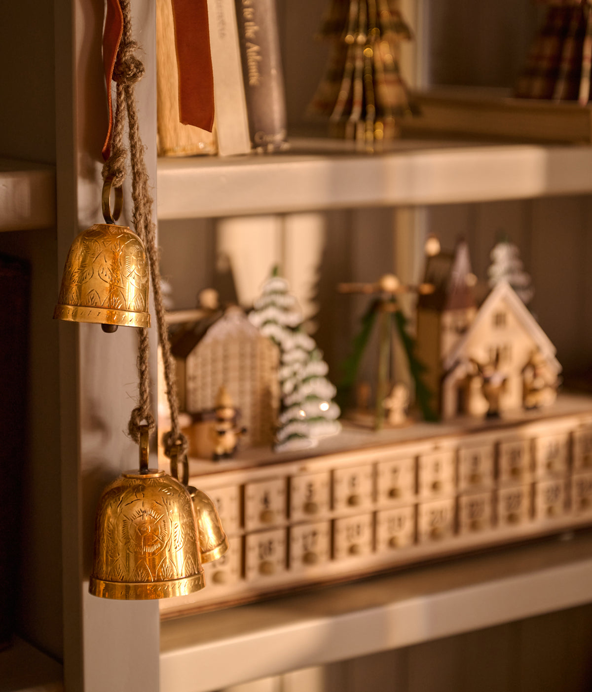 A close-up of a warm, cozy shelf display featuring two hanging brass bells on the left, with a miniature Christmas advent calendar and small house ornaments in the background.